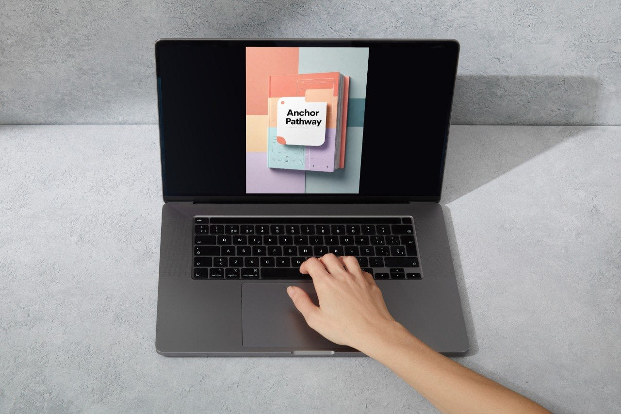 A gray laptop sits open on a light gray surface, with a hand resting on the trackpad. The screen displays a colorful graphic with the text 'Anchor Pathway'.