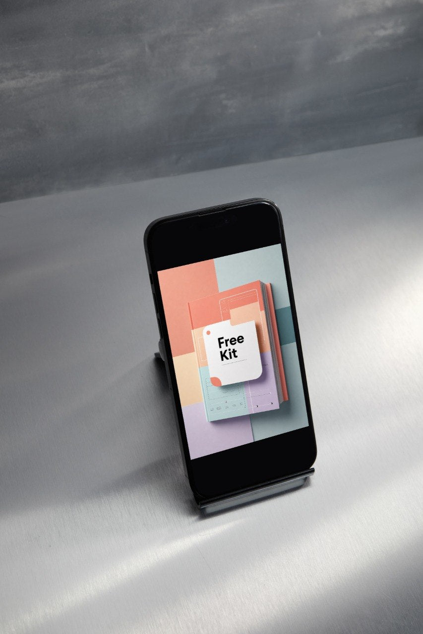 A smartphone displayed upright on a small stand on a light gray surface, with a dark gray textured background. The phone screen shows a colorful app or digital cover labeled 'Free Kit'.