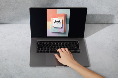 A gray laptop on a light gray surface with a hand resting on the trackpad. The screen displays a colorful graphic with the text 'Spark Course'.